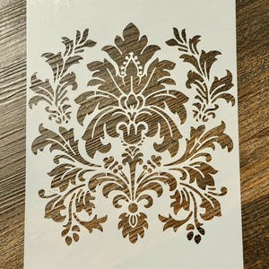 May include: A white rectangular stencil featuring a symmetrical damask pattern with intricate leaf and floral motifs. The design is centered on the sheet, showcasing detailed cutouts that form a repeating organic shape with curved lines.