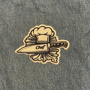 May include: Embroidered patch featuring a chef's hat and a knife. The knife is positioned through a bundle of what appears to be lettuce. The word "Chef" is written on the knife. The patch is white with black outlines and is set against a blue denim background.