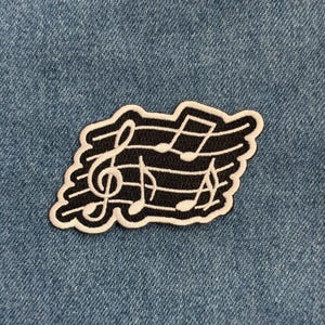 May include: A black and white embroidered patch featuring musical notes and a treble clef. The patch has a black background with white musical symbols and a white border. The patch is placed on a blue denim background.