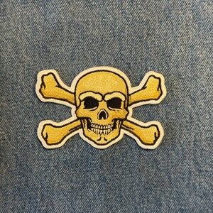May include: Embroidered patch featuring a yellow skull and crossbones design with black eye sockets and a white border. The patch is set against a blue denim background, suitable for jackets, bags, or other apparel.