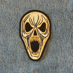 May include: Embroidered patch featuring a screaming skull design with a black border. The skull has a beige color with black eye sockets and mouth. The patch is placed on a blue denim background.