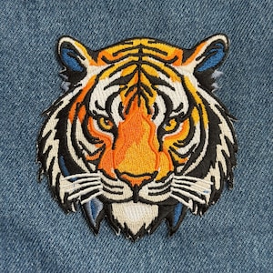 May include: Embroidered tiger head patch with orange, black, white, and blue details. The patch features a detailed design of a tiger's face, with a focus on the eyes and stripes. The patch is likely for clothing or accessories.