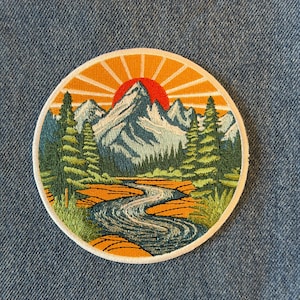 May include: Embroidered patch featuring a mountain landscape with a river, trees, and a setting sun. The patch is round with a white border. The mountains are blue and white, the sun is red and orange, and the trees are green.