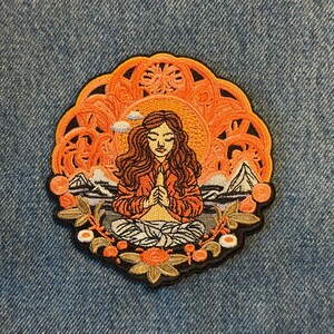May include: Embroidered patch featuring a meditating figure with long brown hair, hands clasped in prayer. The patch has an orange and yellow mandala-like background with mountains and clouds. The patch is bordered with black.
