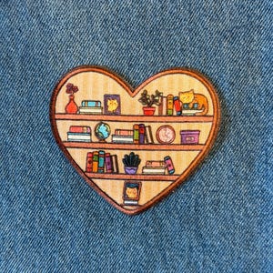 May include: A heart-shaped wooden pin featuring a bookshelf design. The shelves are filled with books, a globe, a cat, and plants. The pin has a brown border and is set against a blue denim background.