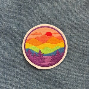 May include: A round embroidered patch featuring a colorful mountain landscape with a setting sun. The design includes layers of mountains in shades of orange, yellow, and purple, with green and blue accents. The patch is bordered with a white edge.
