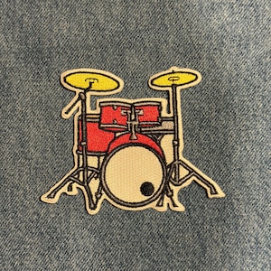 May include: Embroidered patch featuring a cartoon drum set. The drum set has red drums, yellow cymbals, and a black and white design. The patch is on a blue denim background.