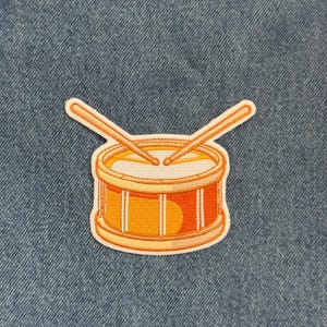 May include: Embroidered patch featuring a cartoon drum with drumsticks. The drum is orange and yellow with a white interior. The patch has a white border and is set against a blue denim background.