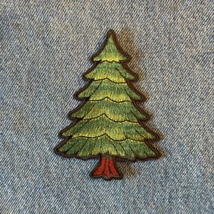 May include: Embroidered patch of a green Christmas tree with a brown outline and a red trunk. The tree patch is on a blue denim background. The patch is approximately 2.5 inches tall.