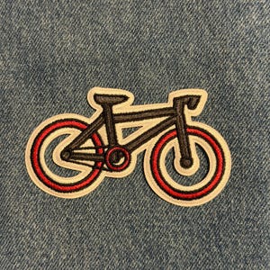 May include: Embroidered patch featuring a bicycle design. The bicycle has a black frame with red and white accents on the wheels. The patch is outlined in white and is set against a blue denim background.