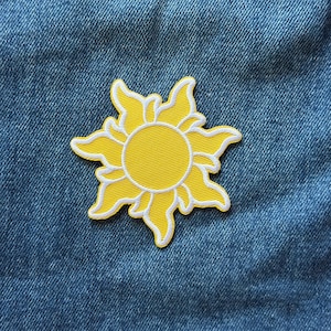 May include: A yellow embroidered patch in the shape of a stylized sun with a circular center and curved, flame-like rays extending outward. The design features a white border outlining the yellow sun shape, set against a blue denim fabric background.