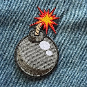 May include: An embroidered patch featuring a round, dark gray bomb with a lit fuse. The fuse is topped with a bright yellow and red starburst explosion. The bomb has two white circular highlights on its surface, creating a glossy appearance.
