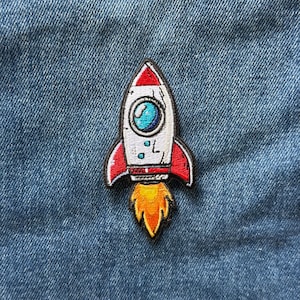 May include: An embroidered patch shaped like a rocket ship with a white body, red fins, and a blue porthole window. The rocket features a bright orange and yellow flame exhaust at the base, all outlined in black thread against a blue denim fabric.