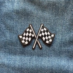 May include: An embroidered patch featuring two crossed checkered racing flags with black and white squares. The patch is stitched onto a blue denim fabric background, displaying a classic racing motif with clean, defined edges and borders.