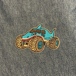 May include: Embroidered patch featuring a shark-shaped monster truck. The truck is primarily light blue with brown and red accents. The tires are tan with a detailed tread pattern. The patch is on a denim background.