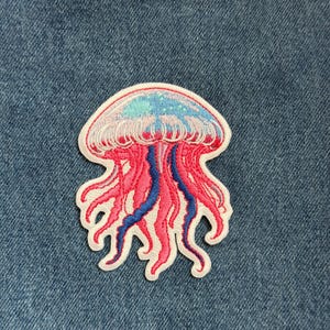 May include: Embroidered jellyfish patch with a white border. The jellyfish features a pink body, blue and pink tentacles, and a light blue and pink top. The patch is set against a blue denim background.