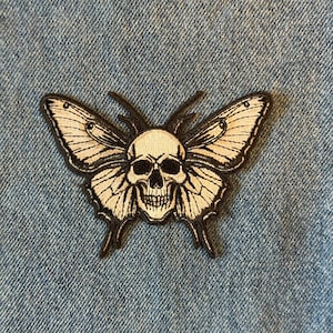 May include: Embroidered patch featuring a butterfly with a skull design. The patch has a black outline with cream-colored wings and a skull in the center. The patch is on a blue denim background.