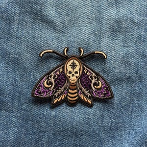 May include: An embroidered patch shaped like a moth with a skull design on its thorax. The moth features purple and tan wings with intricate swirling patterns, a tan striped abdomen, and two curved antennae, all outlined in dark brown thread.