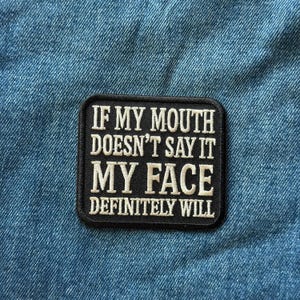 May include: A rectangular black embroidered patch with rounded corners featuring white stitched text that reads: IF MY MOUTH DOESN'T SAY IT MY FACE DEFINITELY WILL. The patch has a thick black border and is placed on a blue denim fabric background.