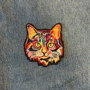 May include: Embroidered cat patch with a colorful design. The cat's face features yellow, red, orange, and blue hues, with a black border. The patch is placed on a denim background.