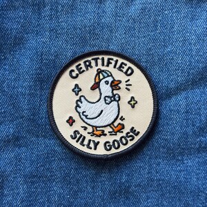 May include: A circular embroidered patch with a cream-colored background and a black border. The patch features a white goose wearing a blue and yellow cap and a bow tie, surrounded by small colorful stars. The text reads CERTIFIED SILLY GOOSE.