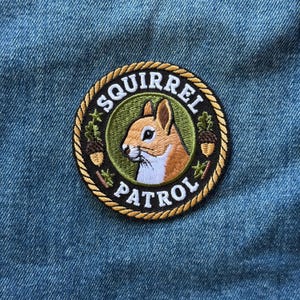 May include: A circular embroidered patch featuring a brown and white squirrel centered on a green background. The border is a gold rope design with the text SQUIRREL PATROL in white letters. Two small brown acorns and green leaves are visible.
