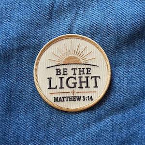 May include: A circular embroidered patch with a cream-colored background and a gold-colored border. The design features a stylized sun rising over a horizon line, with the text "BE THE LIGHT" and "MATTHEW 5:14" embroidered in dark brown thread.