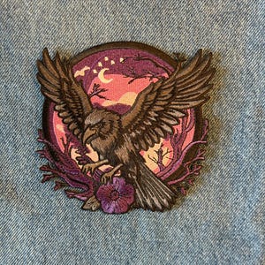 May include: Embroidered patch featuring a detailed illustration of a hawk with outstretched wings. The design includes a night sky with a crescent moon, stars, and stylized trees, set within a circular frame. The color palette includes shades of brown, purple, and pink.