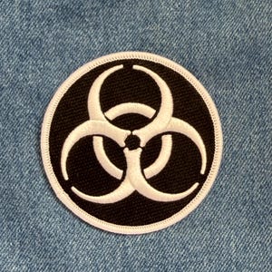 May include: Round black patch with a white biohazard symbol. The symbol is centered on the patch and has three curved arms. The patch has a white stitched border and is set against a blue denim background.