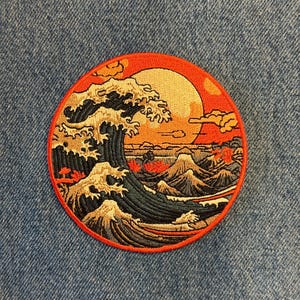May include: Round embroidered patch featuring a Japanese wave design with a red and orange sunset, mountains, and a large wave. The patch has a red border and is on a blue denim background.