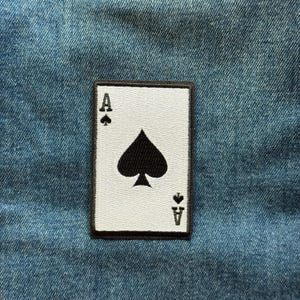 May include: A rectangular embroidered patch featuring the Ace of Spades playing card design. The patch has a white background with a large black spade symbol in the center, and the letter A with a small spade icon in the top left and bottom right.