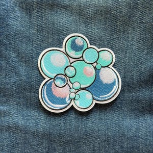 May include: An embroidered patch featuring a cluster of circular bubbles in various shades of blue, teal, and light green. The design includes white highlights on each bubble to create a glossy effect, all outlined with a thick white border.