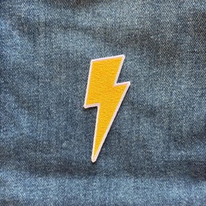 May include: A yellow lightning bolt patch with a white border, featuring a textured embroidered surface. The patch is centered on a blue denim fabric background, showcasing a classic zig-zag shape with sharp points at the top and bottom edges.