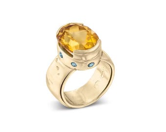 May include: A gold ring featuring a large, oval-cut yellow gemstone. The ring has a textured band and is accented with small, round, light blue gemstones. The ring is set against a white background.