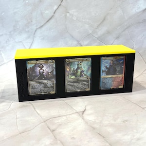 Op de afbeelding: Een zwarte kaarten display case met een felgele bovenkant. De case bevat drie ruilkaarten met kleurrijke illustraties en tekst. De tekst "STRATA LAB" is op de zijkant van de case gedrukt.