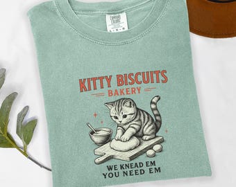May include: A light green t-shirt with the text "KITTY BISCUITS BAKERY" above an illustration of a cat kneading dough. The text "WE KNEAD EM YOU NEED EM" is below the illustration. The shirt is a Comfort Colors brand.