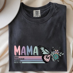 May include: Dark gray t-shirt with the word "MAMA" in pastel colors, a butterfly graphic, and floral accents. Below the word "MAMA" are the words "LOVED, HARDWORKING, SELFLESS, PROTECTIVE". The shirt is made of a soft material.