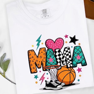 May include: White t-shirt with a colorful graphic design. The word "MAMA" is displayed with various patterns and colors, including leopard print, checkered, and teal. A basketball, hoop, sneakers, and stars complete the design.