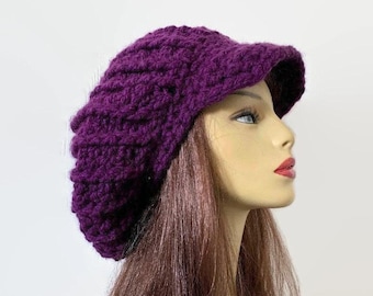 May include: A deep purple, hand-crocheted hat with a visor. The hat is made of thick yarn, with a textured pattern. The visor is a solid color and is slightly curved. The hat is shown on a mannequin head.