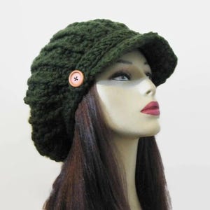May include: A dark green crocheted hat with a brim and a wooden button detail. The hat is a textured knit design and is worn by a mannequin with long brown hair. The hat is a stylish accessory.