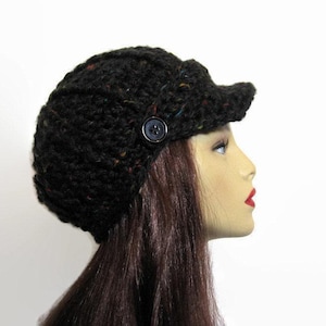 Könnte beinhalten: Schwarze Strick-Newsboy-Cap mit kleinem Schirm und Knopfdetail. Der Hut ist aus einem strukturierten Garn mit Farbflecken gefertigt. Die Mütze wird auf einem Schaufensterkopf gezeigt.