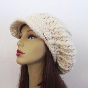 May include: Cream-colored, crocheted hat with a visor and a silver-tone buckle. The hat has a textured, chunky knit design. The brim is slightly curved. The hat is shown on a mannequin head.