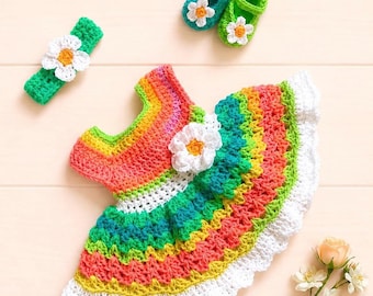 May include: A crochet baby dress featuring horizontal rainbow stripes in pink, orange, yellow, green, and teal, accented with a white flower. The set includes a matching green headband and two small green booties, each adorned with a white flower.