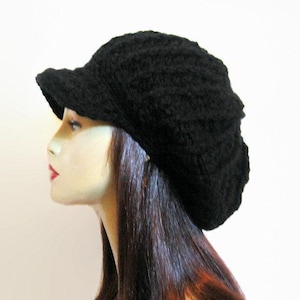 May include: A black knitted hat with a small peak. The hat is made of a textured material, possibly wool. The hat is worn by a mannequin with long, dark hair and red lipstick.