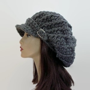 May include: A dark gray, crocheted hat with a small brim and a silver-colored buckle detail. The hat has a textured, woven appearance and is shown from a side profile.