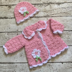 May include: A handmade pink crochet baby set, including a jacket and a matching hat. The set features white scalloped edges and a white flower applique with green leaves. The jacket has long sleeves and button closures.