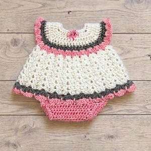 Puede incluir: Un pelele de beb&eacute; tejido a ganchillo a mano con un cuerpo de color crema y cuello redondo, acentuado por una franja gris oscuro y un borde de volantes rosa. La parte inferior es de punto rosa s&oacute;lido con un borde festoneado y detalles grises.