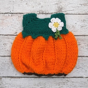 May include: A crochet pumpkin-shaped garment featuring a bright orange body with vertical ribbed texture, a dark green scalloped top section, and a white flower applique with a yellow center attached to the upper right side of the green bodice.