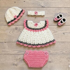 Puede incluir: Un conjunto de ganchillo que incluye un gorro color crema con ribetes rosas y gris oscuro, una diadema a juego con una flor central, un vestido color crema con detalles rosas y gris oscuro, un par de zapatos rosas y un cubrepa&ntilde;al de ganchillo rosa.