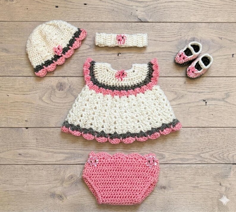 Puede incluir: Un conjunto de ganchillo que incluye un gorro color crema con ribetes rosas y gris oscuro, una diadema a juego con una flor central, un vestido color crema con detalles rosas y gris oscuro, un par de zapatos rosas y un cubrepa&ntilde;al de ganchillo rosa.